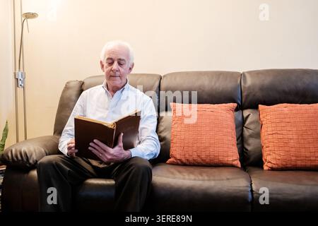 Senior man relaxing on a sofa, reading an old book indoors, enjoying leisure time Stock Photo