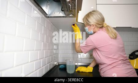 Woman wearing yellow gloves cleans white subway tiles on kitchen wall with sponge near faucet Stock Photo