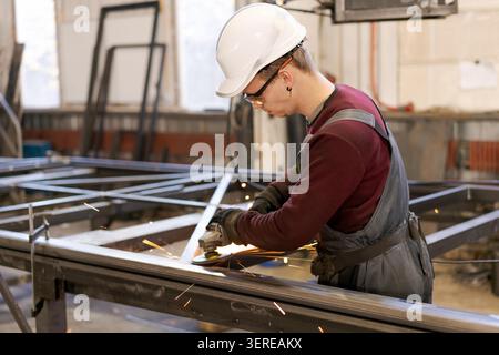 Young tradesman in white hardhat using angle grinder on metal construction with sparks in wide factory shot Stock Photo