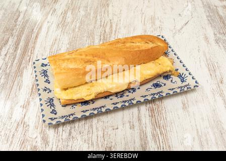 A crusty baguette sandwich filled with thick folded French omelette sits on a blue and white patterned rectangular plate over wood. Stock Photo