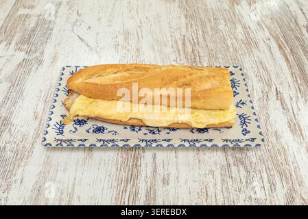 French omelette bocadillo in golden crusty bread served on a decorative blue floral rectangular plate over whitewashed wooden surface. Stock Photo