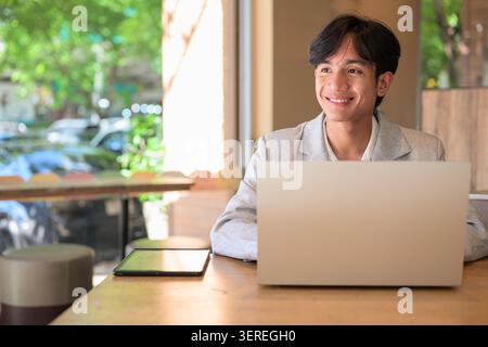 Authentic horizontal lifestyle portrait of a young non-binary Thai Isan professional or student looking away and smiling warmly. Sitting at a wooden t Stock Photo