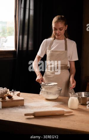 Young woman sifting flour for homemade baking, preparing dough with natural ingredients in a rustic kitchen Stock Photo