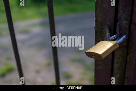 Brass padlock locked on weathered iron latch of rusted gate with vertical metal bars and soft-focus green landscape beyond; shallow depth of field hig Stock Photo