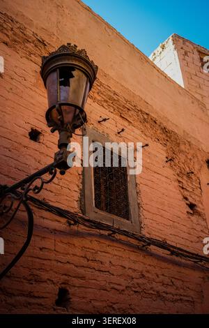 An antique street lamp with intricate metalwork and broken glass is mounted on a textured, reddish-orange plaster wall. Below the lamp, a small window Stock Photo