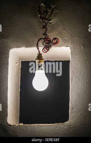 A single incandescent light bulb is illuminated, hanging from tangled red and blue electrical wires. The bulb and its wires are framed by a roughly te Stock Photo