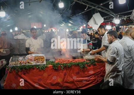 A bustling night market scene unfolds in Marrakech's Djemaa el-Fna square, where several Moroccan chefs are actively preparing and grilling various st Stock Photo