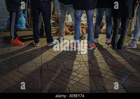 A low-angle view captures the legs and feet of multiple people standing on a dark, geometrically patterned paved surface. Strong, elongated shadows st Stock Photo