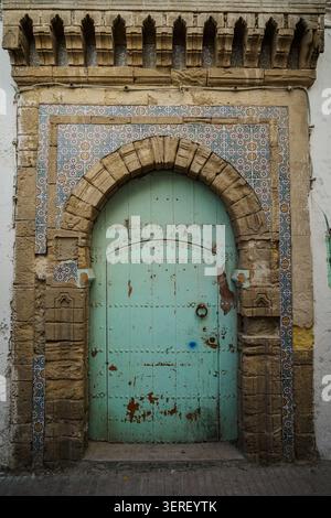 A close-up vertical shot of an aged and weathered wooden door painted in a peeling turquoise hue, featuring a rusted metal handle. The arched doorway Stock Photo