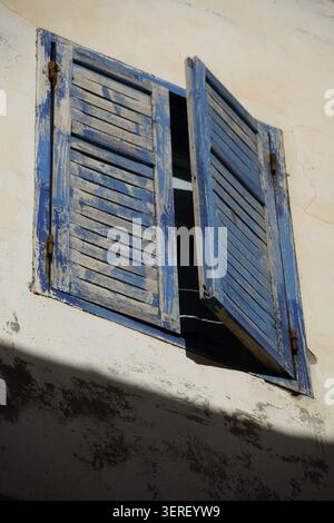 Two distressed blue wooden shutters are seen partially open on an old, textured, light-colored plaster wall under strong sunlight. The shutters show s Stock Photo