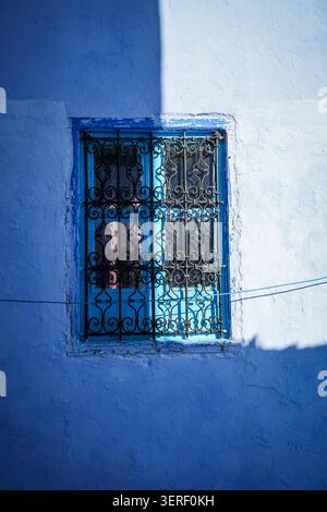 A close-up view captures a vibrantly painted blue wall with a turquoise-framed window, adorned with intricate black wrought iron security bars. Harsh Stock Photo