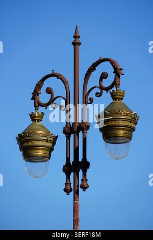 A detailed low-angle shot of an ornate antique street lamp, featuring a rusty brown cast iron pole with decorative scrollwork arms and two brass-color Stock Photo