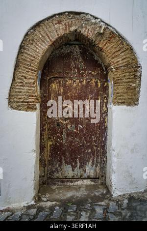 A close-up shot of an ancient, weathered wooden door featuring peeling dark brown and yellow paint, adorned with numerous metal studs and a round door Stock Photo