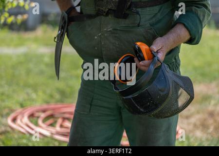 Worker preparing a protective face shield and hearing protection before operating a brush cutter. The image illustrates safety procedures, personal pr Stock Photo