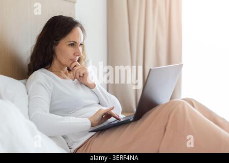 Thoughtful adult woman working on laptop while sitting in bed Stock Photo