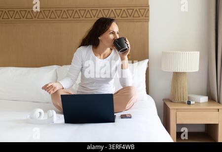 Woman drinking coffee while using laptop on hotel bed, working remotely Stock Photo