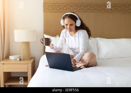 Woman in headphones drinking coffee while working on laptop in hotel room Stock Photo