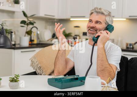 Middle-aged man at home lifts wired landline phone smiles with nostalgia and says warm hello at table on sofa. Mature guy holds handset close shares kind words warm tone and enjoys quick brief call Stock Photo