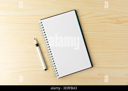 A black marker pen and an open blank lined spiral notebook over a wooden desk. A minimalist flat lay composition representing writing, education Stock Photo