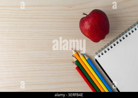 A red apple, colored pencils, and an open blank spiral notebook on a wooden desk. Back to school, education concept. Copy space for text. Stock Photo
