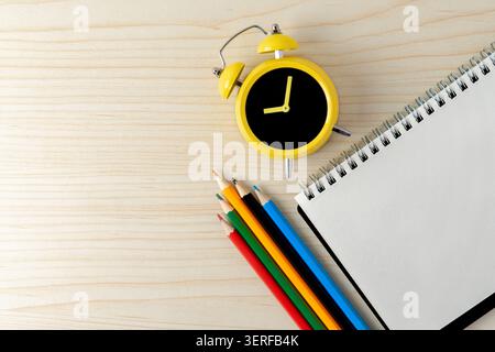 A yellow alarm clock, colored pencils, and a blank open spiral notebook on a wooden desk. Back to school, study schedules, and time management concept Stock Photo