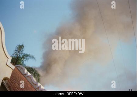A large dark smoke plume billows into a blue sky above a brick building with decorative white trim and palm trees, suggesting a nearby fire. Stock Photo