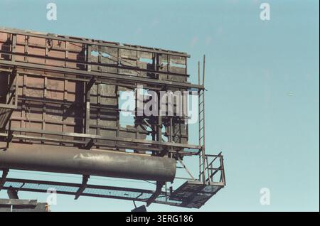 Rusted and broken metal billboard structure with missing panels photographed from below against a clear blue sky, conveying urban decay and neglect. Stock Photo