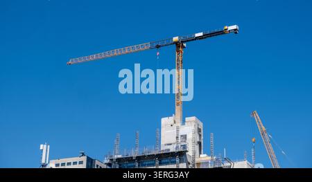 Construction cranes stand over a high-rise work site. Yellow tower crane and mobile crane against a blue sky Stock Photo