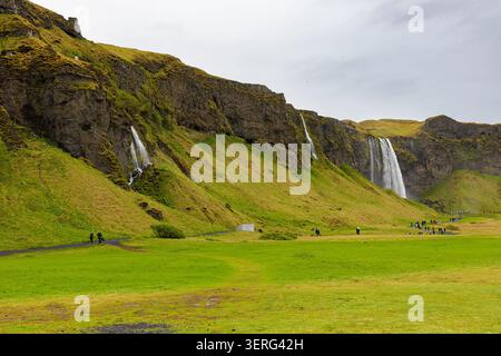 Scenic view of Seljalandsfoss waterfall and smaller cascades along mossy volcanic cliffs with tourists in a vast green meadow in Iceland. Stock Photo