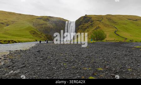 The majestic Skógafoss waterfall rushing down lush green hills with tourists gathered on a volcanic pebble riverbed under an overcast sky in Iceland. Stock Photo