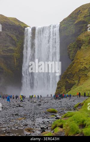 A scenic view of the rushing river flowing from Skógafoss waterfall, with lush green hills under a bright cloudy sky in Iceland. Stock Photo