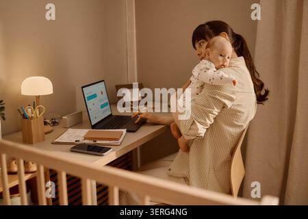 Adult Mother Working on Laptop While Holding Baby at Home Office Stock Photo