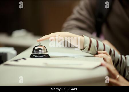Close up of child hand reaching and pressing hotel reception bell while family checking in at front desk. Ideal for travel booking, hospitality service, family vacation marketing Stock Photo