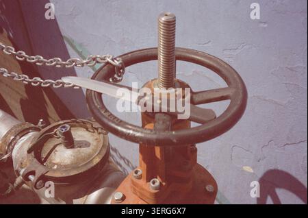 Close-up of an orange industrial gate valve with a circular handwheel, threaded stem, and metal security chain against a weathered concrete wall. Stock Photo