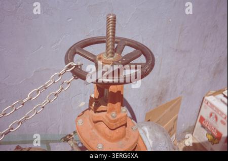 Close-up of a large orange industrial gate valve with black cast iron handwheel and threaded stem, secured with a metal chain against a gray textured Stock Photo