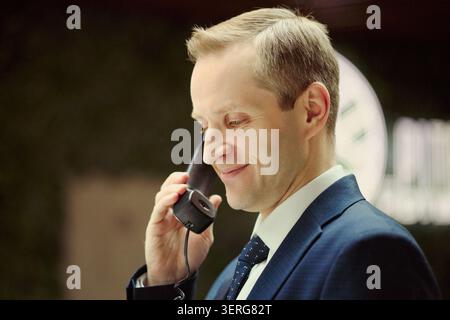 Close up of smiling Caucasian middle aged male concierge speaking on hotel phone, handling guest communication and travel service support. Ideal for hospitality, booking, concierge marketing Stock Photo