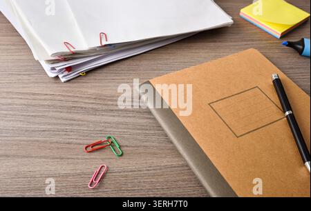Brown notebook, stacked papers, paper clips, sticky notes and pen arranged on a wooden desk, representing office work, documentation and organization. Stock Photo