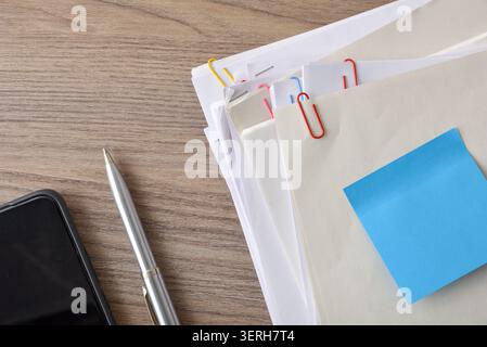 Top view of stacked folders and papers with colorful clips, a silver pen, smartphone edge, and blue sticky note on a wooden desk. Stock Photo