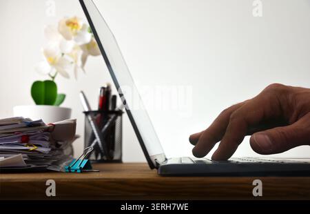 Close side view of a hand typing on a laptop at a desk with paperwork, office supplies, and a plant, representing productivity, administration, and re Stock Photo