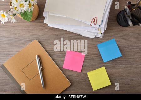 Top view of an organized desk with a notebook, pen, colorful sticky notes, documents, flowers, and office accessories on a wooden surface. Stock Photo