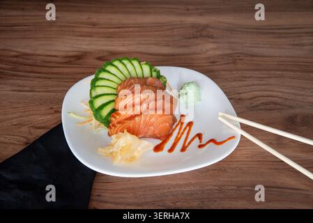 Still life featuring fresh salmon sashimi, sliced cucumbers, wasabi, and ginger on a white plate against a wooden background. Soft, diffused lighting Stock Photo