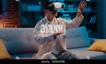 Asian man using modern technology wearing virtual reality helmet playing vr game at home living room at evening. Korean Japanese Chinese male enjoying Stock Photo