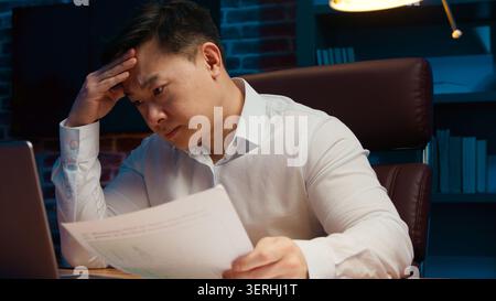 Stressed Asian Korean business man working hard with documents paperwork and laptop difficulties at evening indoors. Frustrated Japanese male Chinese Stock Photo