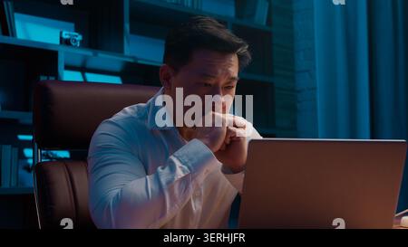 Sleepy exhausted Asian Chinese middle-aged business man thinking with laptop at night at home. Stressed tired Korean Japanese businessman with Stock Photo