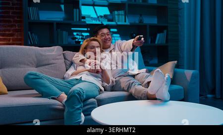 Happy joyful multiracial couple lying on new soft comfortable couch watching tv relax rest in evening at home living room. Multiethnic family Asian Stock Photo