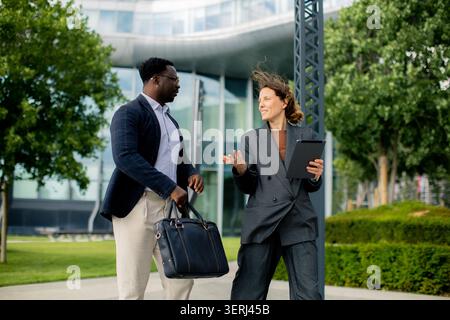In a business setting, two professionals are engaged in conversation near a modern office building. The scene is bright and sunny, and they appear to Stock Photo