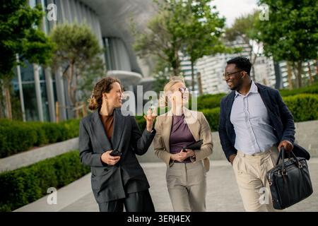 Three people walk together in a park, enjoying conversation. They appear relaxed and engaged. The area has greenery and modern buildings in the backgr Stock Photo