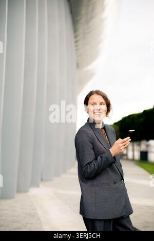 A woman stands outdoors near a modern building. She smiles while holding a phone in her hand. The scene shows green trees and a gray structure in the Stock Photo