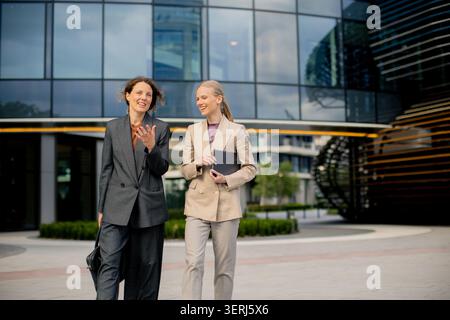 Two women in business suits are walking and chatting outside an office building. They are smiling and holding a tablet while walking on a pathway in t Stock Photo