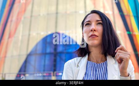 Thoughtful young professional woman standing in front of a modern colorful building Stock Photo